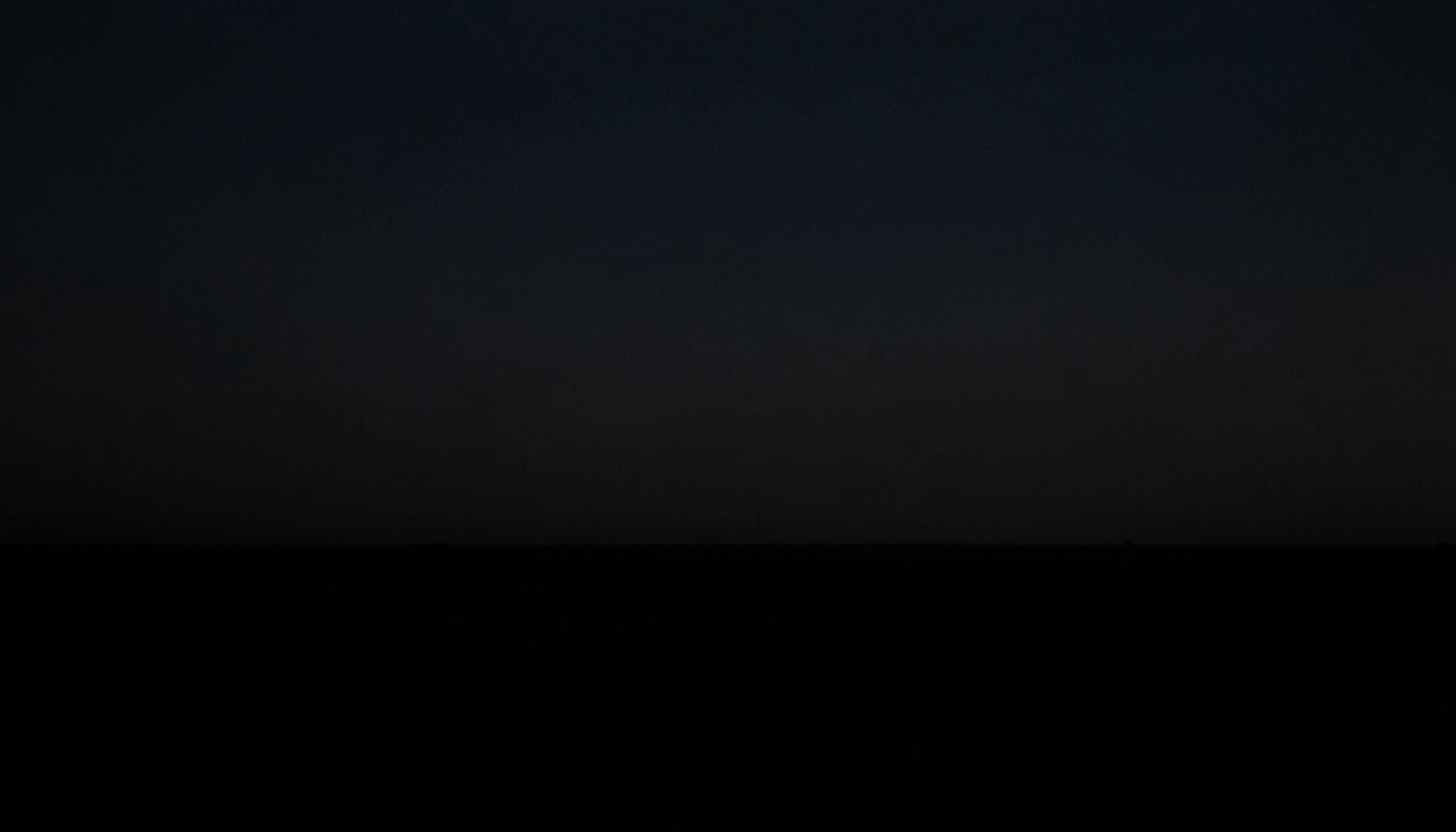 A dark, low-key photograph of a subtle horizon line at dusk, deep charcoal sky meeting near-black land, minimal and serene