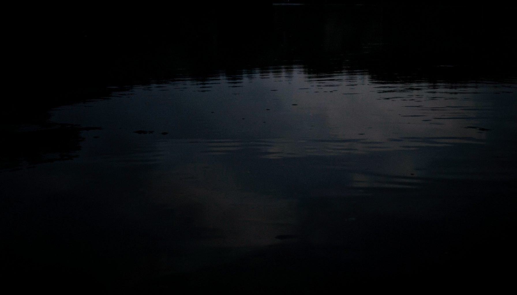 A dark, low-key photograph of still water at twilight, deep charcoal and slate tones with a faint silver reflection on the surface, calm and contemplative mood