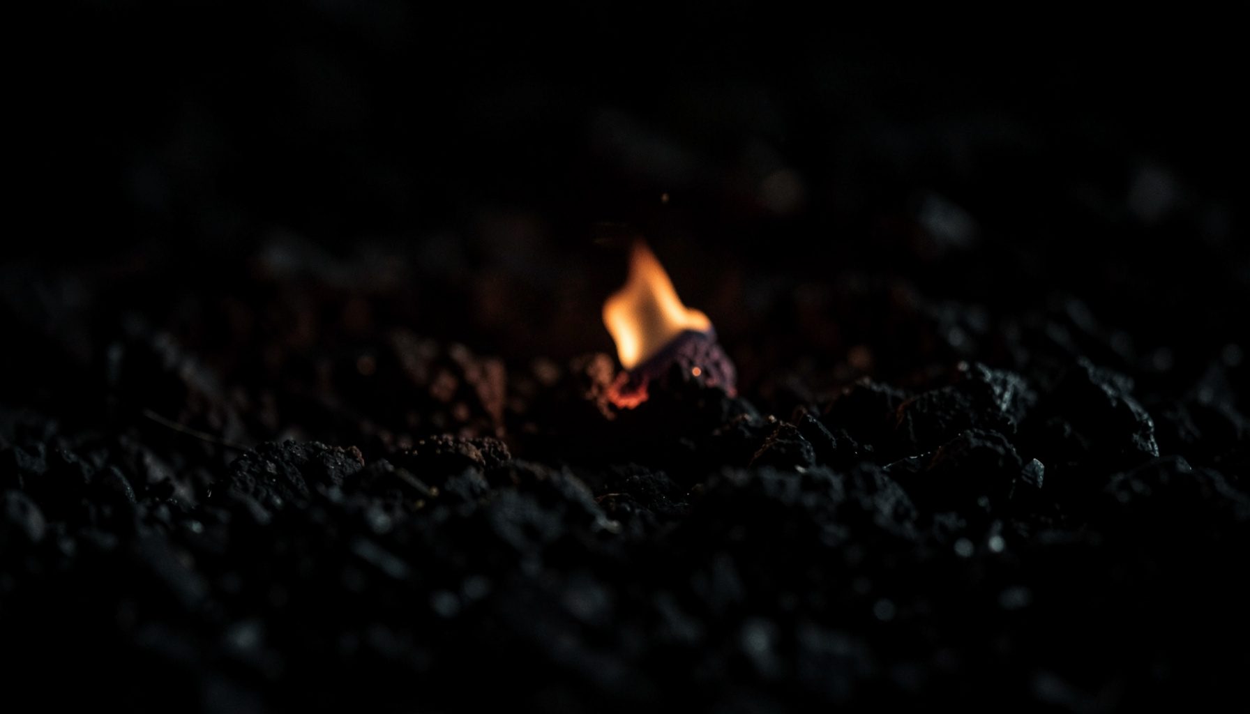A dark, low-key photograph of a single glowing ember resting on charcoal, deep blacks and muted grays with a faint warm orange glow at the center